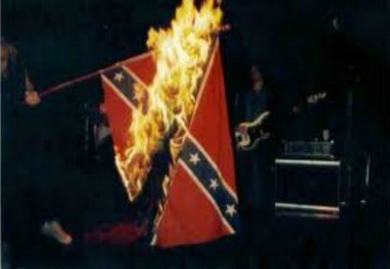 Pimeässä tilassa käsi pitelee etelävaltioiden lippua, joka on liekeissä. Takana erottuu kitaristin siluetti. Kuva ei ole Mustasta Pispalasta.